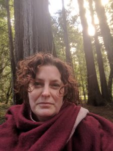 Dr. Margo Wolfe is a white woman with short, curly brown-red hair. She is wearing a crimson shawl and stand outside in a forest with light peeking though behind her.